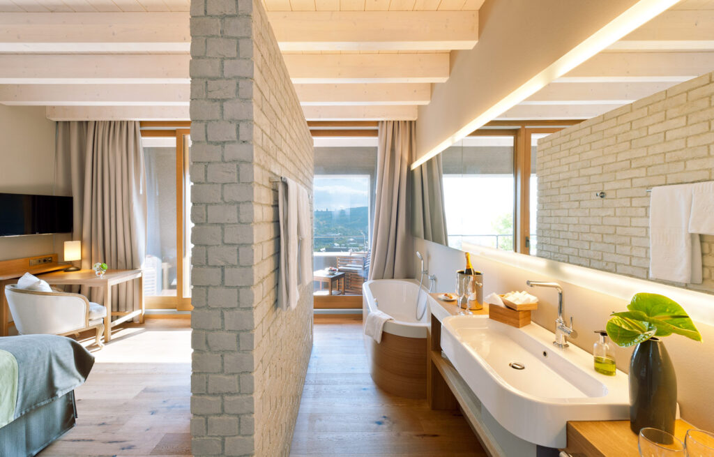 Modernes Hotelzimmer mit offener Gestaltung, freistehender Badewanne und großzügigem Waschbereich. Helle Holzbalken, Natursteinwände und große Fenster mit Blick auf eine Terrasse sorgen für ein luxuriöses Ambiente.