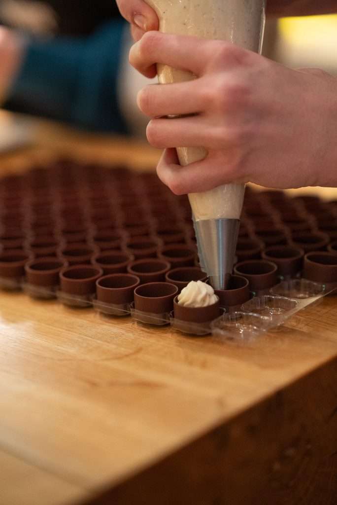 Eine Hand spritzt mit einem Spritzbeutel cremige Füllung in hohle Schokoladenformen auf einer Holzarbeitsplatte.